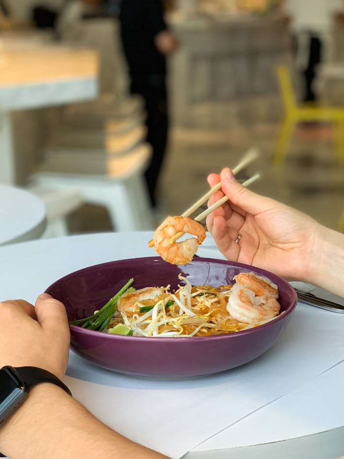 A person enjoying a delicious shrimp noodle meal with chopsticks in a modern dining setting.