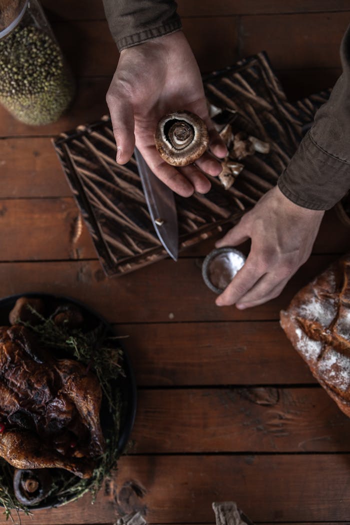 Hands preparing mushrooms for a Thanksgiving dinner, surrounded by turkey and kitchenware on a wooden table.