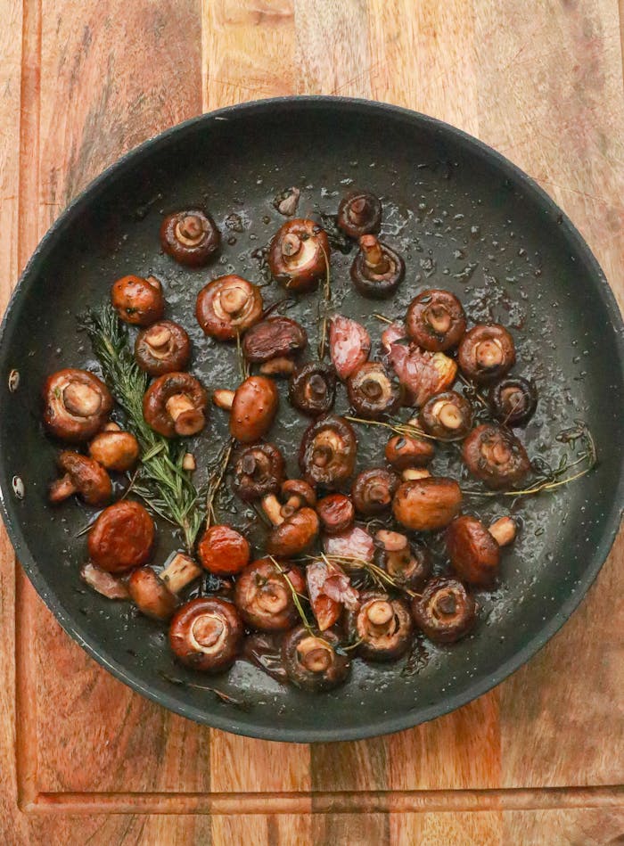 Top view of many little tasty mushrooms in frying pan with green rosemary and garlic placed on wooden table in kitchen